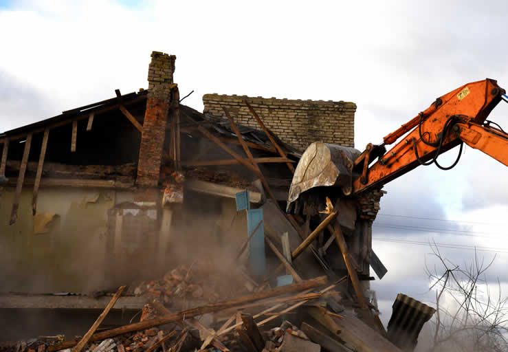 Excavator demolishing damaged house amid rubble